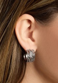 Sølv øreringe i hoop-design med en tekstureret, lagdelt finish, der kombinerer glatte og hamrede overflader, hvilende mod en naturlig øreflip.