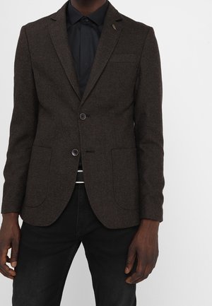Dark brown textured blazer with two buttons and patch pockets worn over a black collared shirt and black pants.