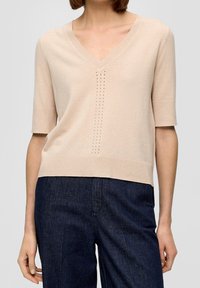 Maglione beige a lavorazione a maglia con scollo a V, maniche corte e un sottile motivo di piccoli fori lungo il centro. Texture morbida, orlo a costine.