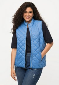 Gilet trapuntato azzurro chiaro con colletto alto, chiusura lampo frontale e tasche laterali. Indossato sopra una camicia scura, abbinato a jeans blu.