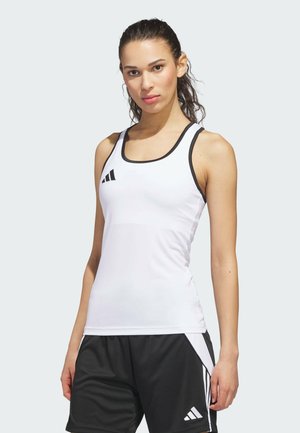 Jeune femme portant un haut de sport blanc sans manches et un short de sport noir avec des rayures blanches, debout devant un fond clair uni.