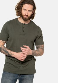 Camiseta Henley verde oliva de manga corta con tapeta de tres botones, textura suave y escote redondo, combinada con pantalones vaqueros azules.