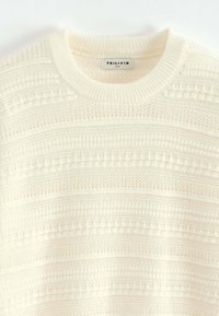 Pull en tricot couleur crème avec un motif texturé, présentant des rayures horizontales et un col rond. L'étiquette indique "FRILIVIN, Taille M."