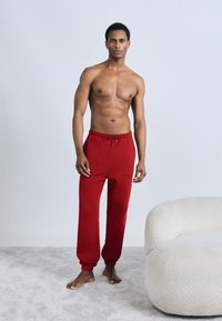 Κόκκινο βαμβακερό παντελόνι φόρμας με ελαστική μέση και κορδόνι, διαθέτει δύο πλαϊνές τσέπες και μανσέτες στα πόδια, φορεμένο ξυπόλητο πάνω σε ένα ανοιχτό χαλί.