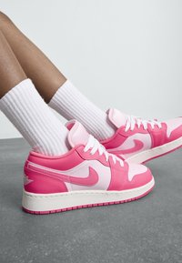 Nogi w białych, prążkowanych skarpetach oraz jasnoróżowych i różowych trampkach Nike odpoczywające na szarym podłożu z jednolitym jasnym tłem.