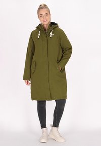 Zelený parkas s kapucí, bílými šňůrkami, bočními kapsami a knoflíkovým zapínáním. Nosený přes tmavé džíny a světlé boty.