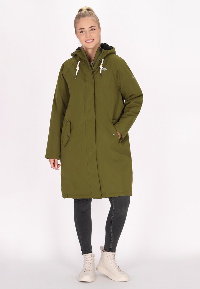 Zelený parkas s kapucí, bílými šňůrkami, bočními kapsami a knoflíkovým zapínáním. Nosený přes tmavé džíny a světlé boty.