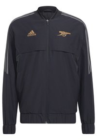 Veste Adidas noire zippée intégrale avec logo Adidas doré sur la poitrine gauche et emblème de canon doré sur la poitrine droite, avec des rayures grises sur les manches.