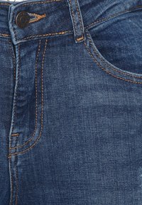 Tecido de denim azul-escuro com costura laranja; apresenta um fecho de botão, passadores de cinto e uma textura subtil que destaca o padrão do tecido.