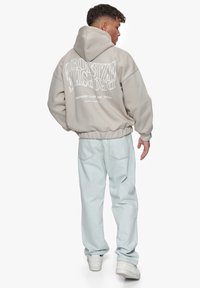 Beige oversized hoodie med hvid trykt tekst på ryggen, parret med lyseblå denim bukser og hvide sneakers. Blød tekstur, afslappet pasform.