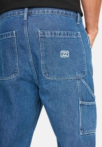 Jeans w kolorze niebieskim średnim. Posiadają dwie tylne kieszenie z białymi przeszyciami oraz naszywką z logo. Prosty krój nogawki z lekko wyblakłą teksturą.