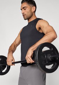 Fekete ujjatlan sportfelső, testhez simuló kialakítással, szürke edzőnadrággal párosítva. Egy személy fekete súlytárcsákkal ellátott súlyzót tart.