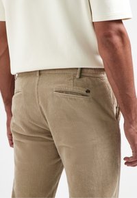 Khaki-gefärbte Cord-Hosen mit vertikaler Ripptextur, ausgestattet mit einer Gesäßtasche, einem gestickten Logo-Akzent und einem Reißverschlussdetail auf der rechten Seite.