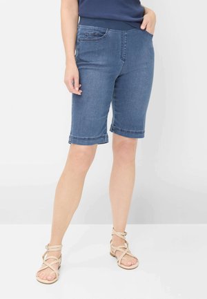 Person iført blå denimshorts til knæene, navy top og beige flade sandaler med remme, der står mod en ensfarvet baggrund.