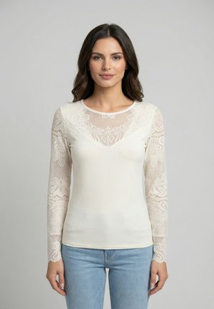 Donna con lunghi capelli scuri che indossa un top bianco in pizzo a maniche lunghe e jeans azzurri, in piedi davanti a uno sfondo grigio uniforme.