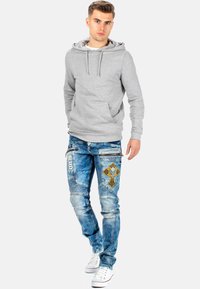Felpa grigia con tasca a marsupio frontale, indossata con jeans blu strappati con un motivo a croce giallo e dettagli con zip, abbinata a sneakers bianche.