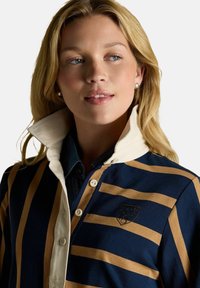 Blonde woman with pearl earrings wearing a navy and tan striped rugby shirt with white collar, looking slightly to the side.