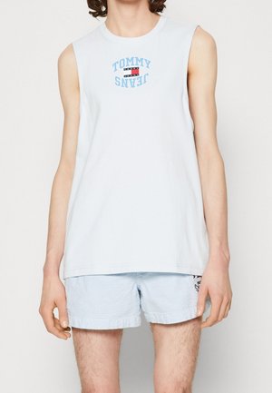 Biały top bez rękawów z logo "Tommy Jeans" w kolorze niebieskim i czerwonym, zestawiony z jasnoniebieskimi i białymi szortami w paski na stojącej osobie.
