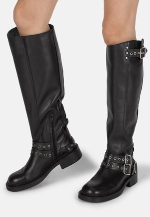 Botas de cuero negro hasta la rodilla con cremalleras laterales, tachuelas metálicas, doble hebilla y cinturones. Tacones gruesos y suelas texturizadas.