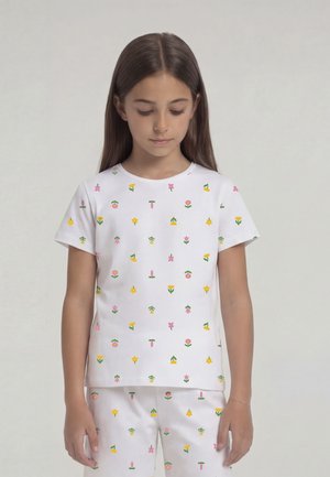 Tüdruk, kes kannab valget lühikeste varrukatega topi ja pükse väikeste värviliste lillemustritega, vaatab alla tavalise valge tausta ees.