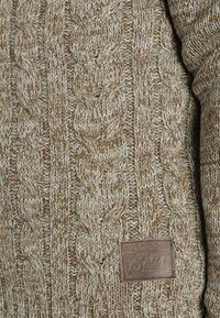 Striktrøje med kabelmønster i beige og creme. Har en struktureret overflade og et brunt lædermærke på kanten.