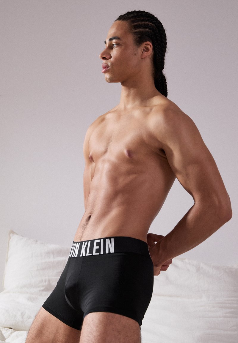Schwarze Baumwoll-Boxershorts mit einem elastischen Bund, auf dem das Logo "CALVIN KLEIN" in Weiß zu sehen ist. Modell von der Seite vor neutralem Hintergrund gezeigt.