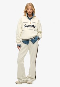 Sweater met ronde hals in ivoor met het "Superdry" logo, gecombineerd met een blauwe jeansshirt en crèmekleurige losse broek met een zijstreepdetail.