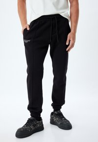Svarta joggingsbyxor i mjukt tyg, med dragsnodd i midjan, sidofickor och diskret logobroderi. Åtsittande mudd vid anklarna.