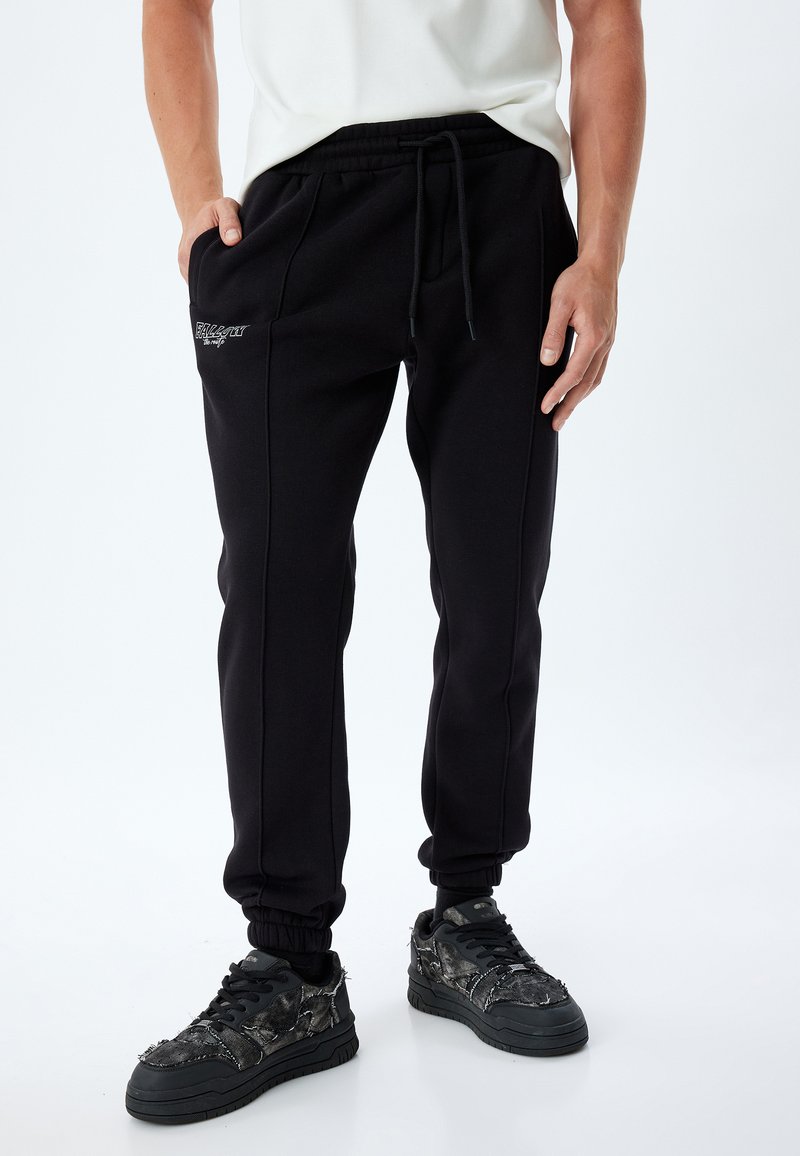 Svarta joggingsbyxor i mjukt tyg, med dragsnodd i midjan, sidofickor och diskret logobroderi. Åtsittande mudd vid anklarna.