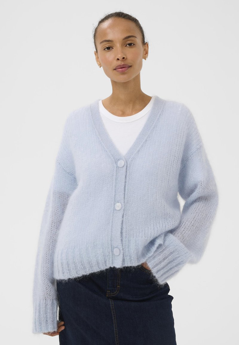 Cardigan bleu clair en tissu tricoté doux. Comprend un col en V, une fermeture à boutons et des poignets côtelés. Porté par-dessus un t-shirt blanc.