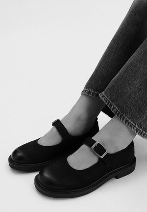 Pies con zapatos Mary Jane de cuero negro con hebillas plateadas, combinados con vaqueros oscuros, sobre un fondo liso.