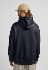 Schwarzer Kapuzenpullover mit glatter Textur, Rippenbündchen und Kängurutasche. Getragen mit einer hellbeigen Kappe und beigen Hosen.