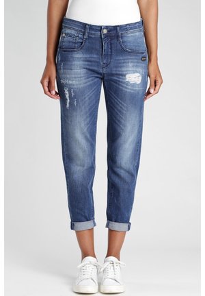 Femme portant un jean bleu déchiré avec revers et des baskets blanches, debout devant un fond uni, mains détendues le long du corps.