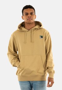 Sudadera beige hecha de una tela suave, con un bolsillo frontal, capucha con cordones y un pequeño parche de logo de outdoor en el lado izquierdo del pecho.