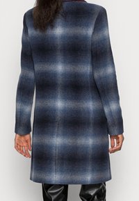 Vrouw in een knielang blauw en zwart gegrafeerd plaid jasje, staand met ontspannen handen langs de zijden, met de rug naar de kijker gekeerd.