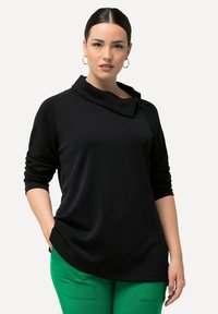Černý top s dlouhým rukávem, cowl krkem a zipovými detaily. Vyrobeno z hladkého, elastického materiálu. V kombinaci se zelenými kalhotami.