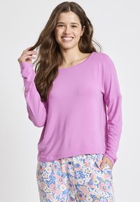 Langärmliges, locker sitzendes Hemd aus leuchtendem pinkfarbenem Stoff, runder Ausschnitt und mit glatter Textur. Getragen mit floral gemusterten Hosen.