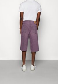 Lila Jeansshorts mit geradem Bein, zwei Gesäßtaschen und einem geprägten Lederpatch am Bund. Getragen mit weißen Sneakers.