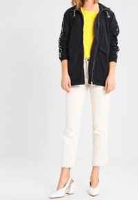 Czarna lekka kurtka z troczkami, w zestawieniu z jasną żółtą bluzką, białymi krótkimi spodniami dżinsowymi i białymi butami na wysokim obcasie.