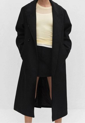 Person mit einem knielangen schwarzen Mantel über einem blassgelben Hemd und einem schwarzen Rock, steht mit den Händen in den Manteltaschen vor einem schlichten Hintergrund.