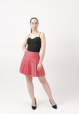 Kobieta stojąca pewnie, ubrana w czarny top bez rękawów, plisowaną spódnicę w czerwono-biały wzór oraz czarne szpilki, na jednolitym białym tle.