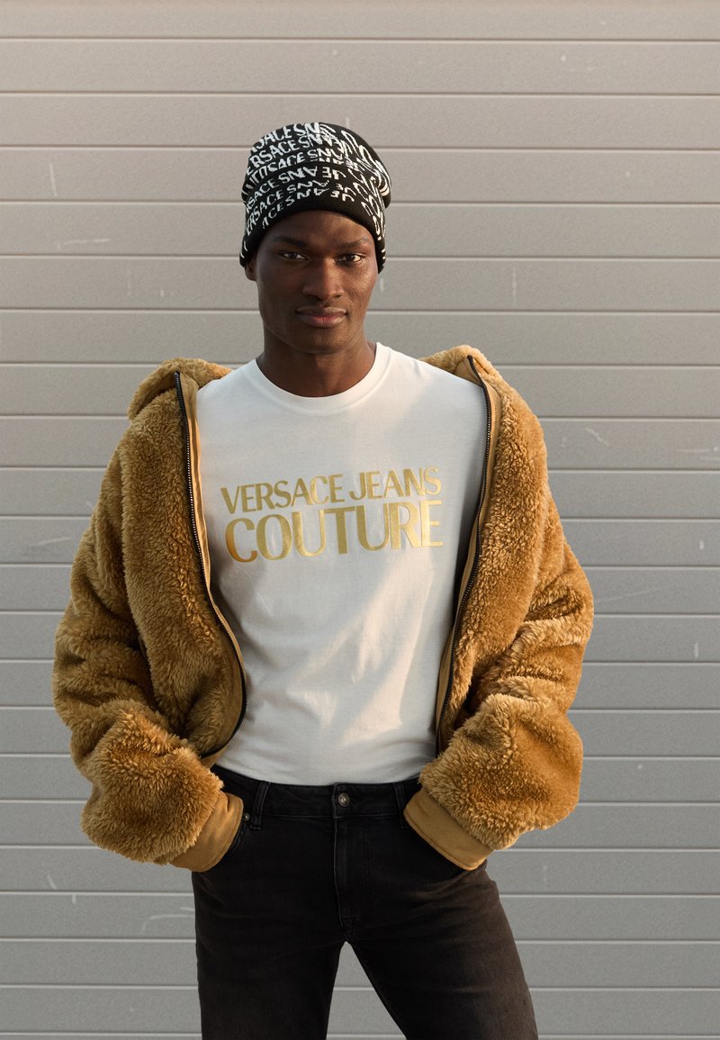 Brūna mākslīgās kažokādas jaka ar rāvējslēdzēju virs baltas T-krekla ar zelta burtiem. Melnas džinsu bikses un melna cepure ar baltu teksta rakstiem.