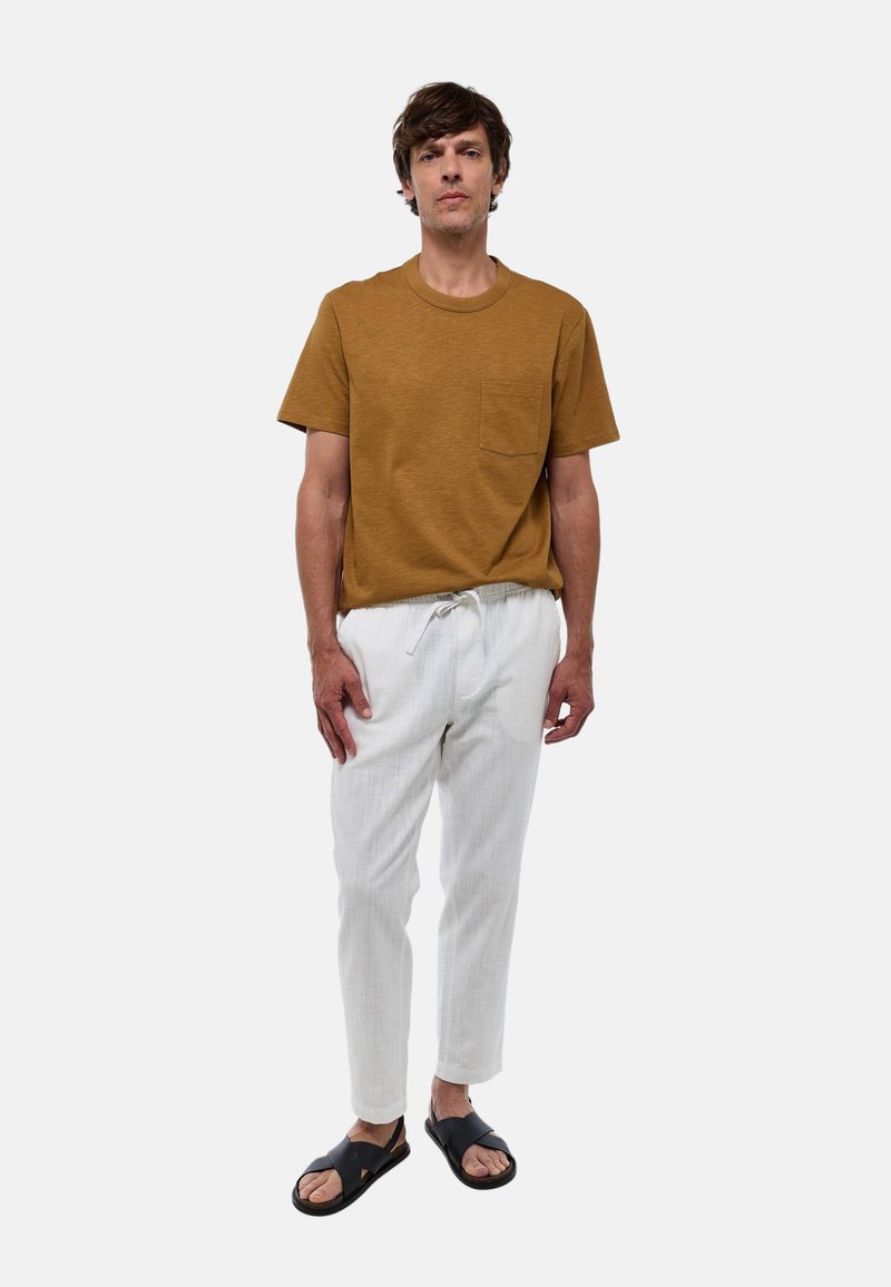 T-shirt marron à manches courtes avec une poche sur la poitrine, associé à un pantalon blanc à cordon de serrage texturé et des sandales noires.
