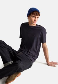 Mladý muž v modré čepici, tmavém tričku, džínách a hnědých botách sedí na podlaze s neutrálním výrazem.