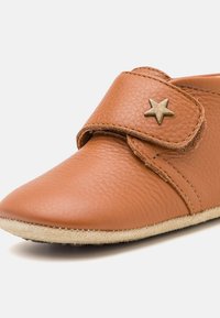 Zapato de cuero marrón con punta redonda y textura suave, presenta una correa de velcro adornada con un detalle de estrella dorada. La suela es ligera y flexible.