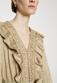 Μπεζ φόρεμα με ρυπανόμενο λαιμό και περίπλοκες λεπτομέρειες από δαντέλα. Λείος υφάσματος με διακριτικά σχέδια, που αναδεικνύει μια V-σχήμα μπροστά.