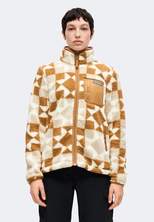 Persona con una chaqueta de forro polar beige y marrón con estampado, cuello alto, cremallera frontal y bolsillo en el pecho, de pie frente a un fondo liso.
