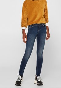 Jeans ajustados azul oscuro con una textura suave, complementados con un suéter de canalé mostaza. El conjunto presenta un diseño casual y zapatillas deportivas.