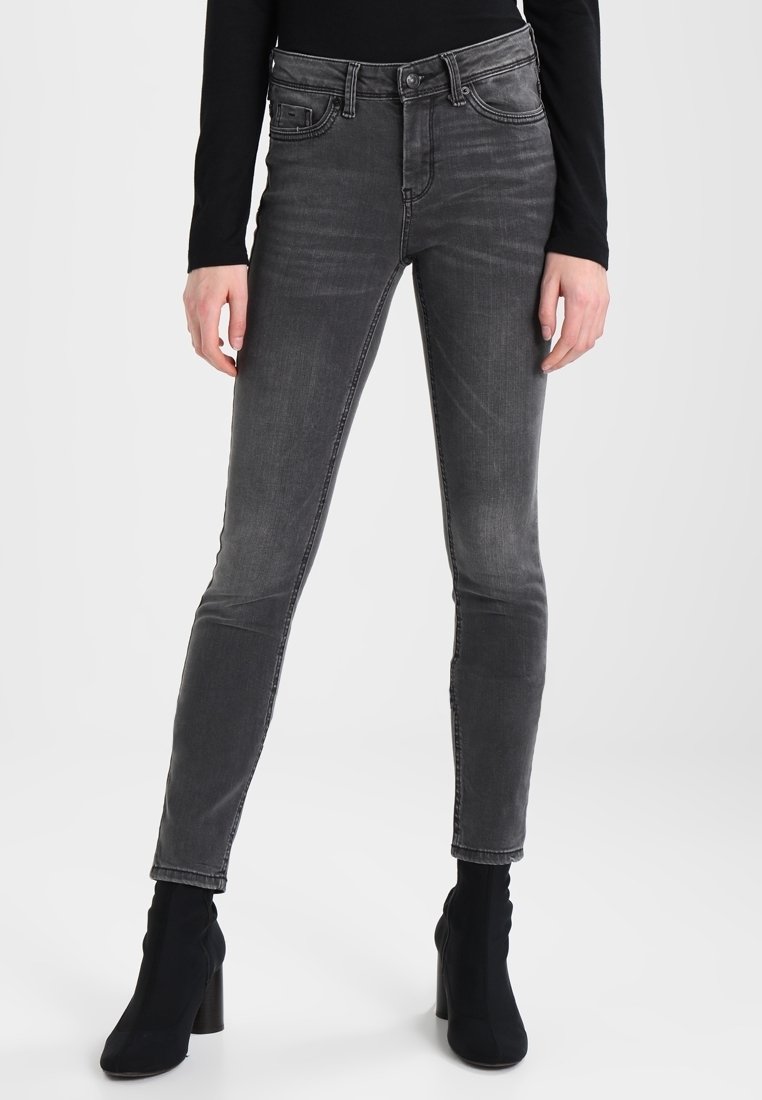 Personne portant un jean skinny noir délavé, un haut noir à manches longues et des bottines noires à talons, debout les mains détendues.