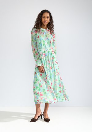 Femme debout dans une robe midi fleurie vert menthe à manches longues et des chaussures slingback marron à talons bas sur un sol blanc.